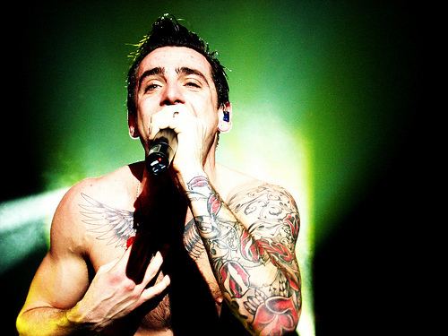 Jacob Hoggard is singing, his left hand holding a microphone, his right hand on his chest, a tattoo cross and wing on his chest, and full sleeve tattoo on his left arm, wearing an earpiece.