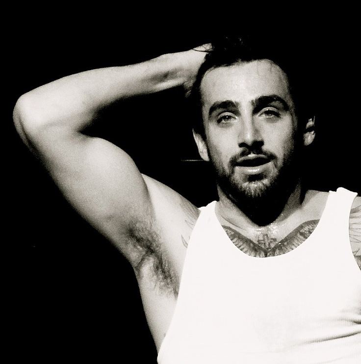 Jacob Hoggard is serious, mouth half opened, has black hair, a beard, and mustache, right hand up showing his armpit with hair, a tattoo of cross above a wing on his chest, and wearing a white tank top.