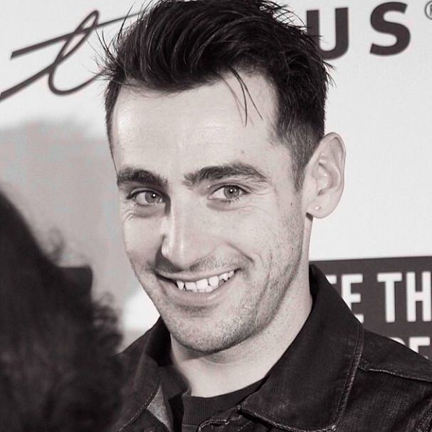 Jacob Hoggard is smiling, has black hair, a beard, and a mustache, wearing an earring on his left ear, and a black shirt under a black jacket.