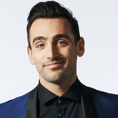 Jacob Hoggard is smiling, has black hair, a beard and mustache, wearing black long sleeves under a black and blue suit.
