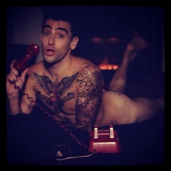 Jacob Hoggard is serious, mouth half opened, has black hair, naked, right hand holding the red telephone, has tattoos on his chest and left shoulder.