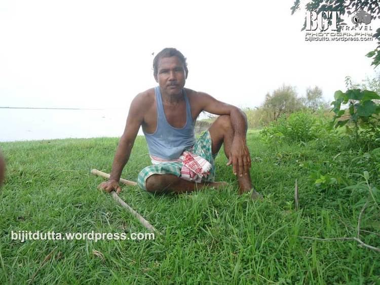 Jadav Payeng sitting on the grass and holding a stick with a serious face and mustache while wearing a blue sando, green and white checkered shorts, and a red and white cloth wrapped around his waist