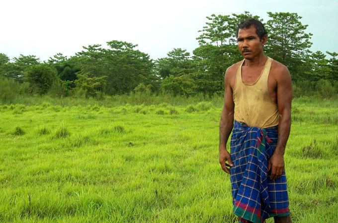 Jadav Payeng looking afar with a serious face, mustache, trees, and green field behind him while he is wearing a brown sando and blue checkered cloth wrapped around his waist