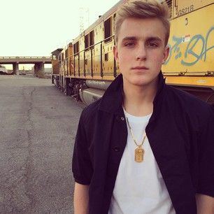 Jake Paul with a poker face while behind him is a yellow train and he is wearing a black jacket, white t-shirt, and necklace