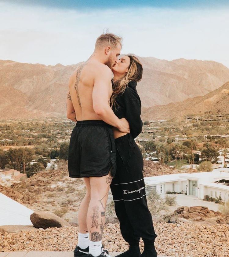 Jake Paul and Julia Rose kissing each other in a place with an overview of the mountain while Jake wearing black shorts, white socks and black shoes