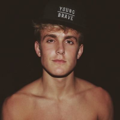 Jake Paul with a tight-lipped smile while wearing a black cap with a "young brave" print on the front