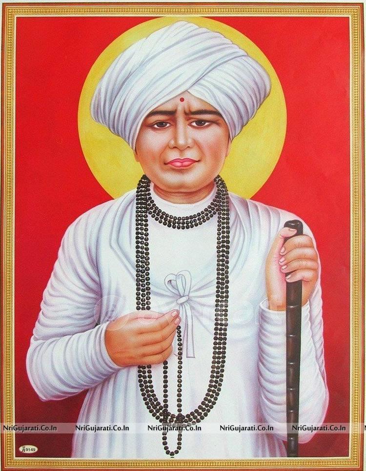 Jalaram Jay holding a wooden stick and a necklace while wearing a white turban, white long sleeves, and a black beaded necklace