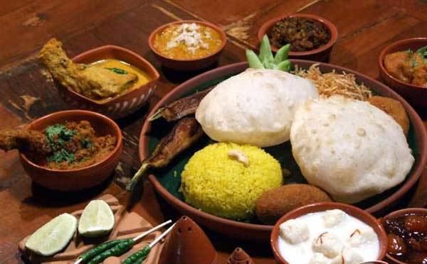 A famous food of west bengal of Jamai Shashthi restaurant, at the bottom left is a side dish lemon and green chilis on, from left a bowl of fried meat in a sauce, 2nd from left is a box bowl of a fried meat in a sauce, 3rd is a bowl of orange sauce with coconut shreds, 4th is a bowl of meat , 5th a bowl of mean in orange sauce, in the middle is a plate of yellow rice fried fish cut cucumber, beansprout, a fried in a ball shape, the bottom right is a Keema doi bora next to it a bowl with dark sauce.