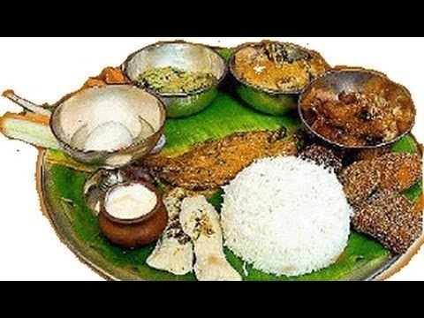 A bengali festival food of Jamai Shashthi restaurant, in a large plate with banana leaf, a white rice with  pot of white sauce, fried fish in the middle, a cup with 2 eggs and a bowl of white meats, a bowl of yellow sauce, a brown meat  with coconut and a bwon of meat in brown inside a stainless bowl.