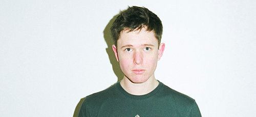 In a white background, James Blake is serious, standing, has brown hair wearing  a green shirt.