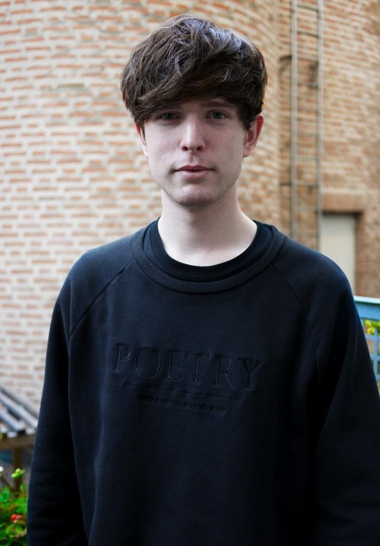 James Blake is serious, standing in front of a tall brick building, has brown hair wearing a black shirt under a black sweater.