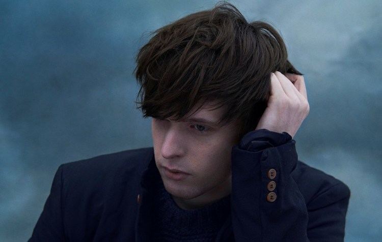 In a cloudy blue-gray background James Blake is serious, looking down to his right with his left hand in his hair, has brown hair wearing a black shirt under a black coat.