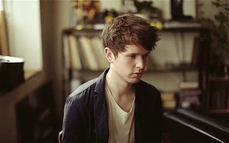In a room with white walls and a brown book shelf at the back filled with books, James Blake is serious, sitting with brown hair wearing a cream shirt under a black coat.