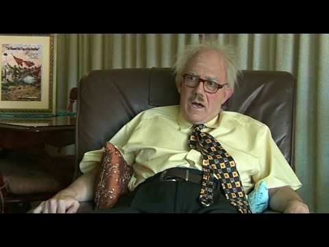 James F. Blake sitting on a couch with two pillows between his arms, with a mustache and gray hair and wearing a yellow shirt, necktie, and black pants with a brown belt