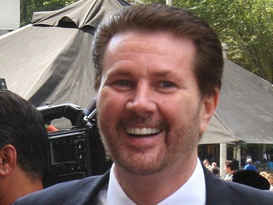 James Galante smiling with mustache and beard while wearing a black coat and white long sleeves
