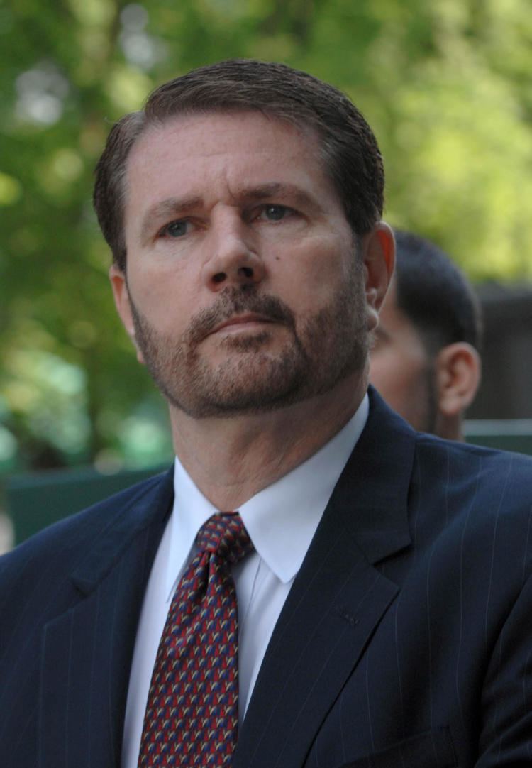 James Galante with mustache and beard while wearing a black striped coat, white long sleeves, and maroon and blue necktie