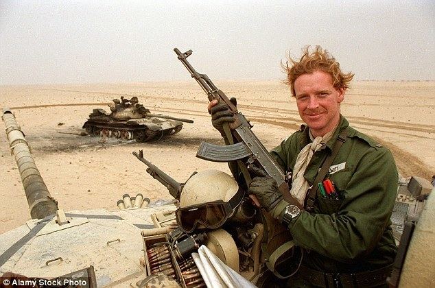 In a desert lands, has brown tank at the back, in front James Hewitt is smiling, standing on top of a tank along with a brown helmet and ammunitions, with his hands holding an ak75 rifle, has brown wavy hair wearing a white scarf a black watch dark olive green gloves and a dark olive green military uniform.