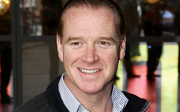James Hewitt is smiling, standing in front of a glass mirror window, has brown hair, wearing a white striped polo under a black jacket.