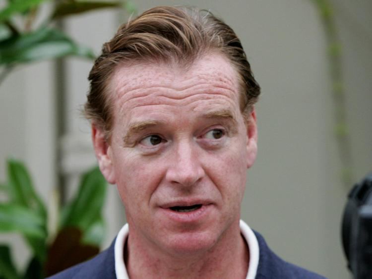 James Hewitt is serious, speaking mouth half open, looking to his left, has brown hair wearing a white polo under a blue coat.