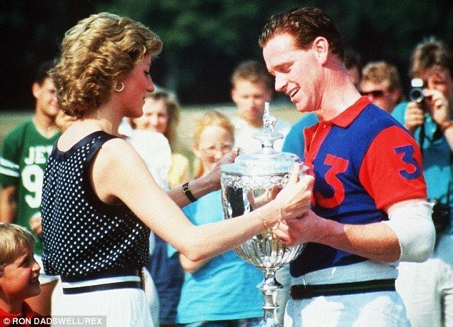 In a field with trees and audience at the back, in front from left, is a boy standing looking up, has blond hair wearing a red polo, in the middle, Princess Diana is serious, standing holding a large silver trophy, has blond short hair wearing a black watch, sleeveless black polka dot top, and white shorts, at the right, James Hewitt is smiling, looking at the trophy, standing holding it with both hands, has brown hair wearing a blue shirt with red collar and sleeve, with number tree on it, a white elbow pad and white pants.