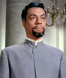 James Lanphier looking afar, with mustache and beard, and wearing gray long sleeves in a scene from the 1963 film, The Pink Panther