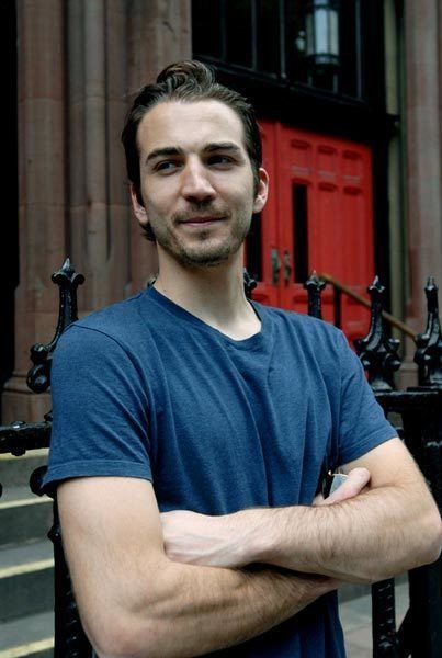 Jamie Johnson with a tight-lipped smile while his arms crossed and wearing a blue t-shirt