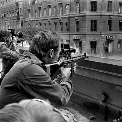 Press photographers holding a camera while taking shots and wearing a coat and police snipers holding a rifle wearing a coat lie side by side on a roof opposite the Kreditbanken bank on Norrmalmstorg square in Stockholm on Aug. 24, 1973.