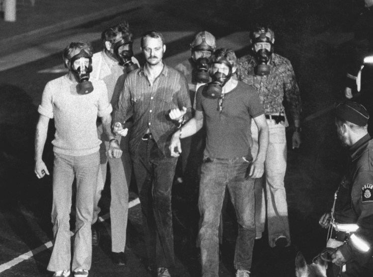 Jan-Erik Olsson (center with no gas masks) seriously looking while in handcuffs his both hands to another two men wearing shirts and pants and with gas masks and 3 other men at their back wearing polo shirts and pants and with gas masks, also a police officer at their left corner looking at them wearing a police cap and police uniform, from a file photo taken on August 23, 1973, at the Kreditbanken bank on Norrmalmstorg square in Stockholm. Jan-Erik has a mustache wearing a polo long sleeves and pants with a black belt