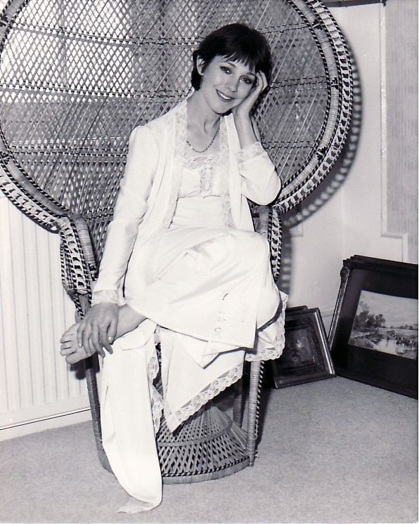 In black and white, Jan Francis is smiling, sitting in a rattan chair in a room with paintings on the floor behind her, has black short hair, wearing a necklace, white sleeping dress under a white robe.