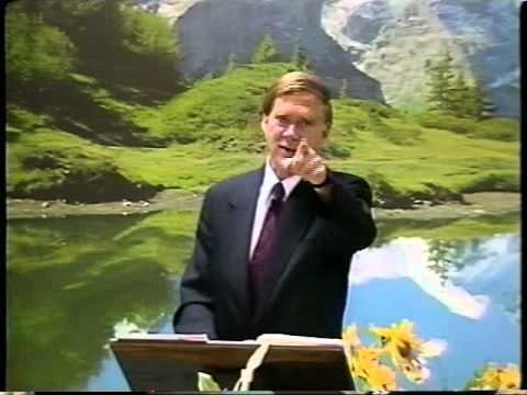 Jan Marcussen giving a speech with hand gestures while wearing a black coat, white long sleeves, and violet necktie