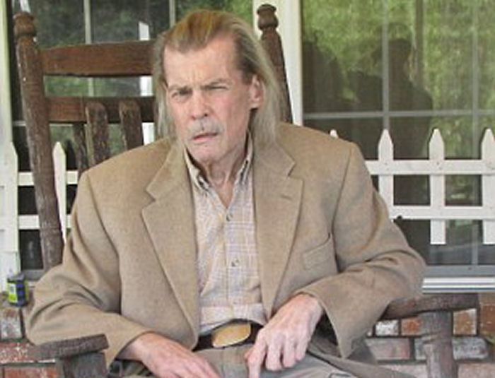 Jan-Michael Vincent sitting on the chair, with mustache and long hair, while wearing a beige coat, pants, light brown long sleeve, and black belt
