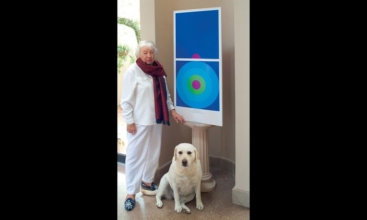 Jane Davis Doggett with a serious face while standing beside her graphic art and a white dog, wearing a white blouse and pants, and a multi-colored scarf.