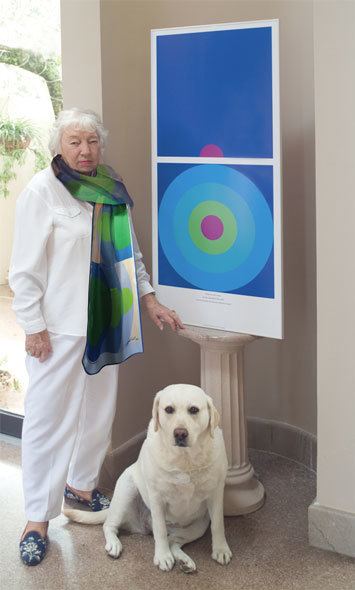 Jane Davis Doggett with a serious face while standing beside her graphic art and a white dog, wearing a white blouse and pants, and a multi-colored scarf.