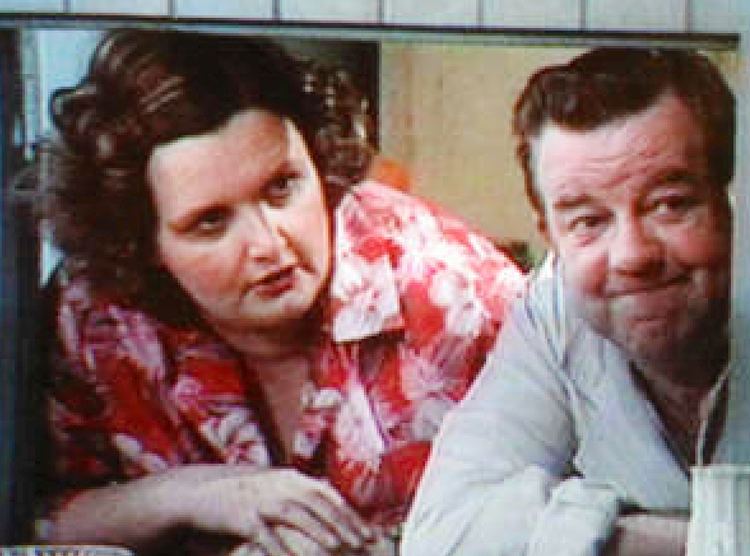 Jane Freeman as Ivy and John Comer as Sid in a 1973 British sitcom TV series, Last of the Summer Wine. Jane with a serious face while talking has brown curly hair, wearing a red Hawaiian-inspired collared polo, while John, smiling forcedly has brown neat-combed hair, wearing a white collared long sleeve.