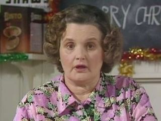 Jane Freeman, talking seriously to someone, an English-born Welsh actress with brown curly short hair and a board and box in a Christmas decoration background. She is wearing a purple floral collared blouse.