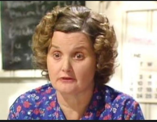 Jane Freeman, talking seriously to someone, is an English-born Welsh actress mostly notably for her role as Ivy in Last of the Summer Wine. She has dark blonde curly short hair with a board and a calendar in the background, wearing a royal blue floral collared blouse.