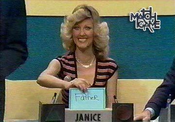 Janice Pennington smiling and holding a paper at the Match Game show with the word "father" written on it and she is wearing a black and pink striped blouse