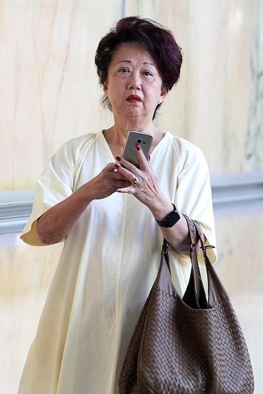 Jannie Chan Siew Lee (Hour Glass Co-founder) is serious while holding her phone with her two hands, has short black hair, and wears a black wristwatch and black ring in her left hand, a white dress, and a brown bag.