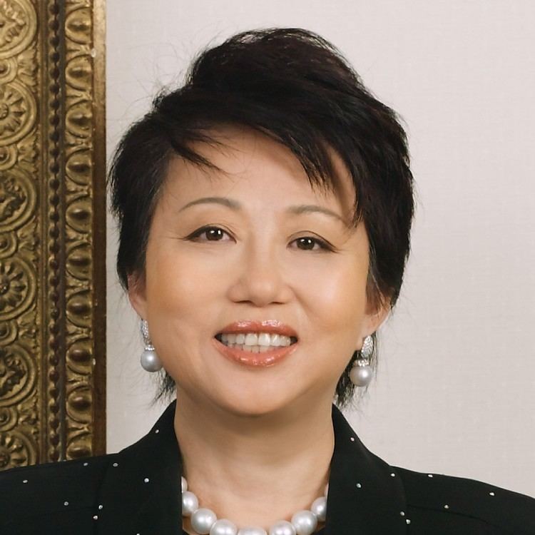 Jannie Chan Siew Lee (Hour Glass Co-founder) is smiling, has black short hair, wearing silver earrings and a necklace, and a black polka dot top. Behind (left) is a Chinese background that is color brown and gold.