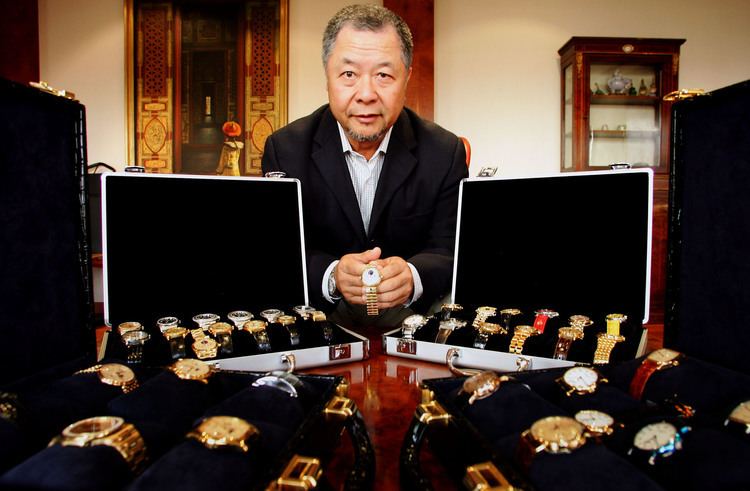 Tay Yun Chwan "Henry" (Chairman/Co-Founder) is sitting down inside The Hour Glass boutique seriously holding a wristwatch with both hands, has black and white hair, and a beard, wearing white long sleeves under a black suit.  In front of him is a table with a gray and black suitcase with a lot of wristwatches inside, it has different styles and colors. Behind him (right) is a glass cabinet with different kinds of stuff inside, on left is a Chinese background with red, black, and gold design with black and gold wine.