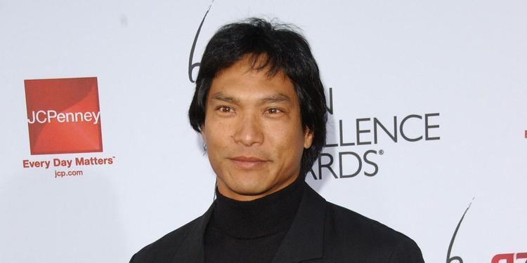 Jason Scott Lee with a tight-lipped smile while wearing a black coat and black turtle neck shirt