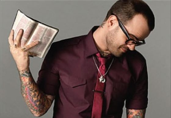 Jay Bakker looking down while holding a bible and wearing maroon polo, red necktie, eyeglasses, and necklace