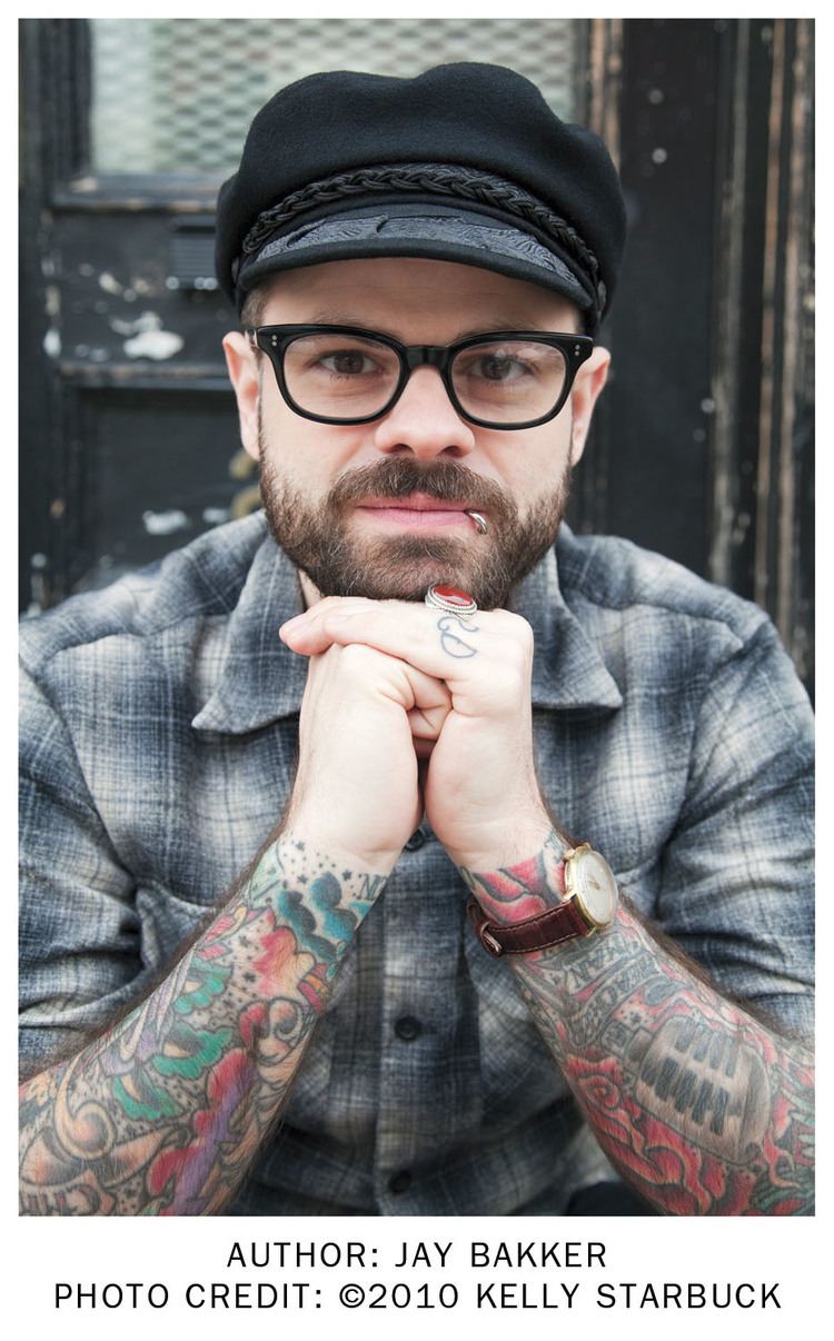Jay Bakker's chin is leaning on his hand, with mustache and beard, while wearing a black cap, eyeglasses, blue and white checkered polo, wristwatch, and lip piercing