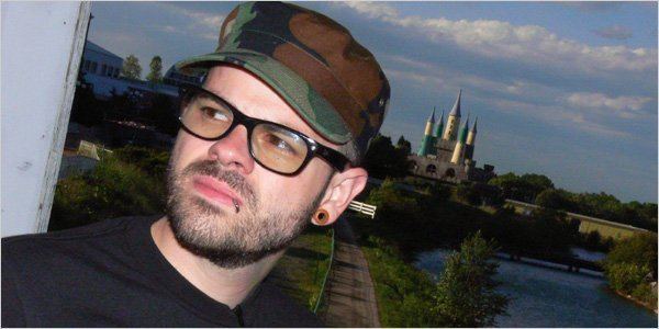 Jay Bakker looking at something while the castle behind him is can be seen and he is wearing a black shirt, eyeglasses, lip & ear piercing, and camouflage cap