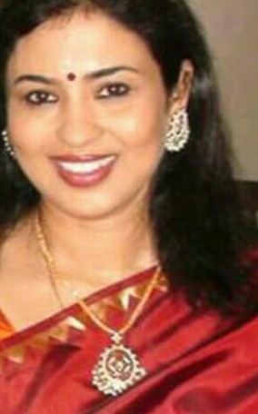 Jayashree with a smiling face, wearing earrings, a necklace, and a red dress.
