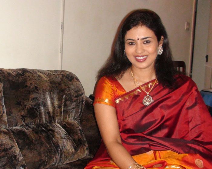 Jayashree with a smiling face, wearing earrings, a necklace, bracelets, and a red and orange dress while sitting on a couch.