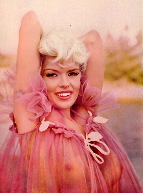 Jayne Mansfield is smiling, has white hair, both hands at the back of her head, wearing a pink see-through breast-showing dress.