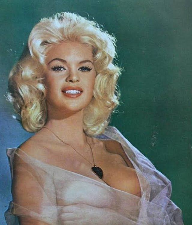 Jayne Mansfield is smiling, has white hair, wearing a black heart necklace, and a cleavage see-through dress.