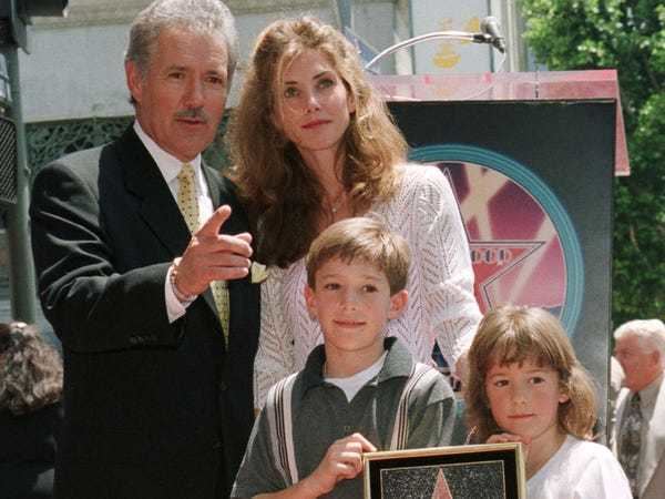 Alex Trebek smiling in a black suit with his wife, Jean Trebek wearing a white dress, with their two children Matthew and Erin.