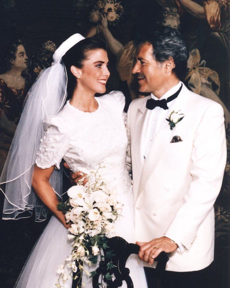 Jean smiling in a white dress and holding a flower bouquet while being held in the waist by her husband, Alex, during their wedding